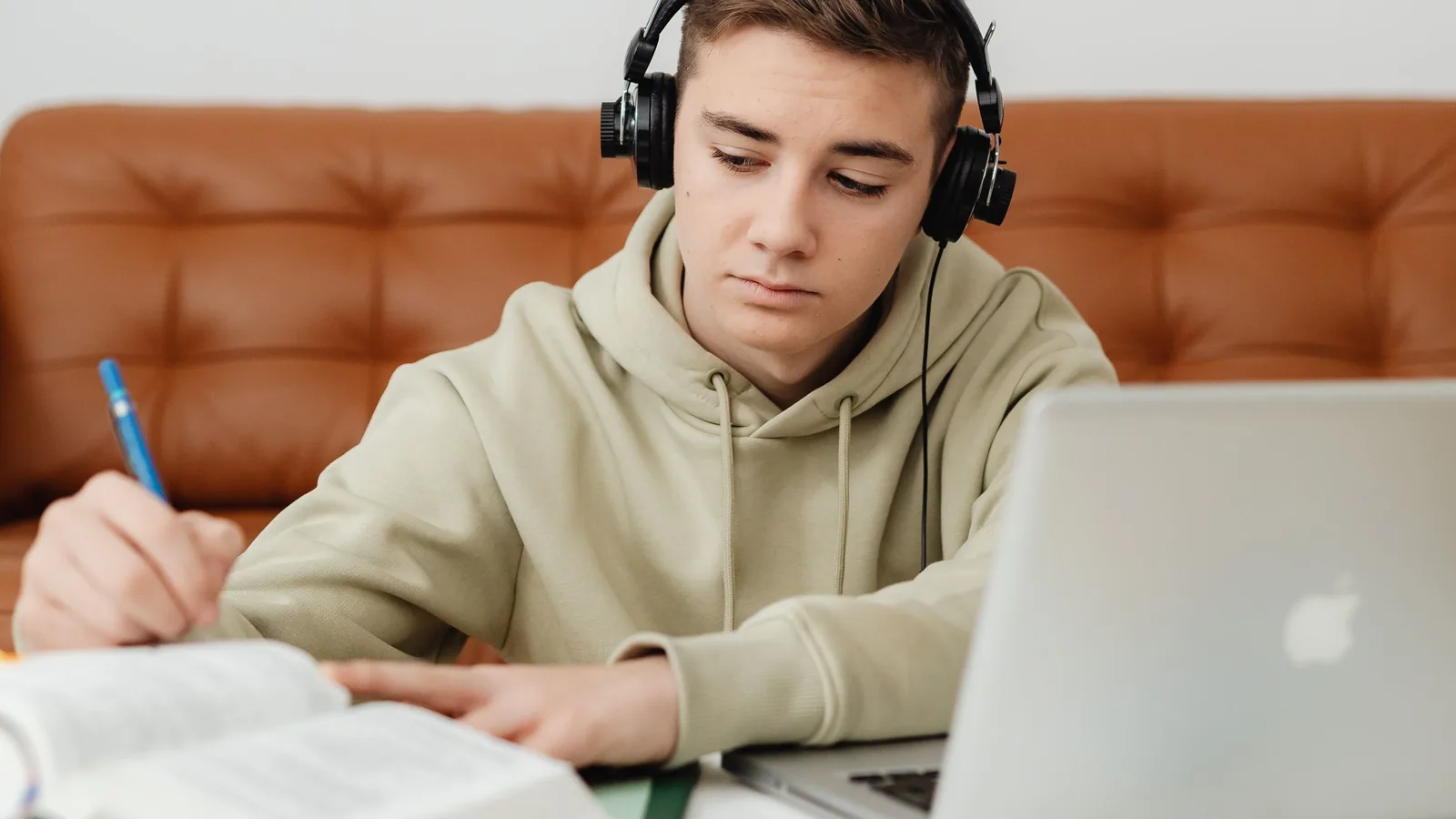 A teenage student in headphones writing notes beside an open book and a laptop at a desk at home